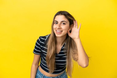 Sarı arka planda izole edilmiş genç beyaz bir kadın. Kulağına elini sürerek bir şeyler dinliyor.