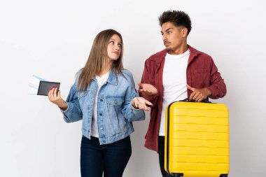 Mavi arka planda izole edilmiş bir bavul taşıyan gezgin çift omuzlarını kaldırırken önemsiz bir jest yapıyor.