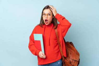 Mavi arka planda, sürpriz bir yüz ifadesiyle izole edilmiş kitapları tutan öğrenci kız.