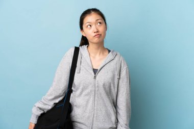 Spor çantalı genç Çinli kadın izole edilmiş mavi arka planda ve yukarı bakıyor