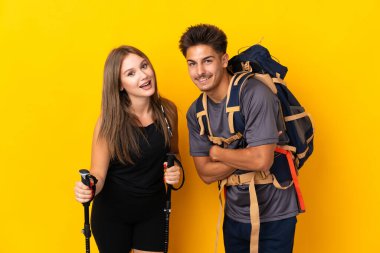 Sarı arka planda izole edilmiş büyük sırt çantalı genç dağcı çift gülümseyerek kollarını kavuşturuyor.