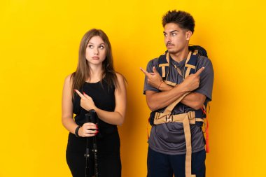 Sarı arka planda izole edilmiş büyük sırt çantalı genç dağcı çifti yan taraftakilerin şüphelerine işaret ediyor.