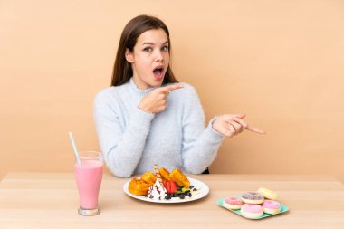 Bej arka planda izole edilmiş waffle yiyen genç kız şaşırmış ve işaret ediyor.