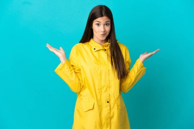 İzole edilmiş mavi arka planda yağmura dayanıklı bir ceket giyen Brezilyalı genç kız ellerini kaldırırken şüpheye düşüyor.
