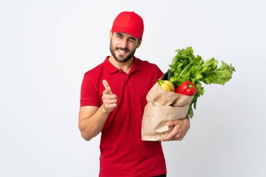 Sakallı genç bir adam elinde sebze dolu bir çanta beyaz arka planda izole edilmiş bir parmak sana.