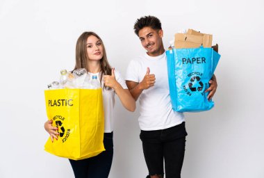 Beyaz arka planda izole edilmiş plastik ve kâğıt dolu bir çanta tutan genç çift başparmak işareti yapıyor.