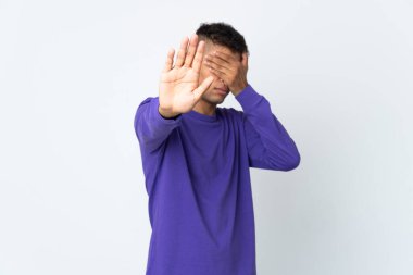 Beyaz arka planda izole edilmiş genç Brezilyalı adam jesti durdurup yüzünü kapatıyor.