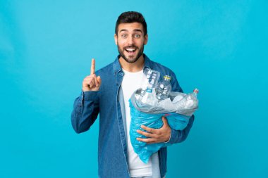 Mavi arka planda izole edilmiş plastik şişelerle dolu bir çanta taşıyan genç beyaz adam harika bir fikre işaret ediyor.