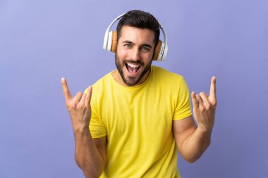 Mor arka planda sakallı yakışıklı genç adam rock hareketleri yaparak müzik dinliyor.