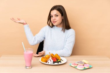 Bej arka planda izole edilmiş waffle yiyen genç kız davet etmek için ellerini yana uzatıyor.