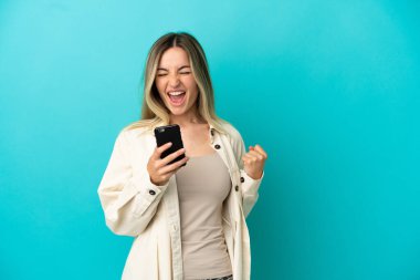 İzole edilmiş mavi arka planı olan genç bir kadın ve telefon zafer pozisyonunda.