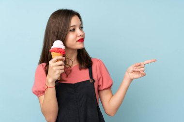 Kornet dondurması tutan genç bir kız mavi arka planda izole edilmiş bir şekilde bir ürünü sunmak için kenarı işaret ediyor.