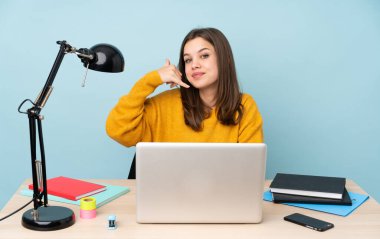 Öğrenci kız, mavi arka planda izole bir şekilde evinde çalışıyor ve telefon jesti yapıyor. Bana geri dönüş işareti ver.