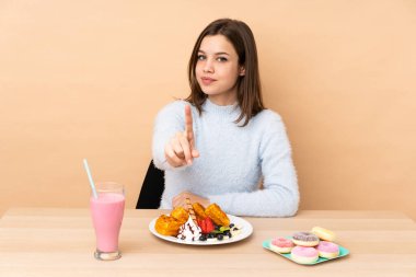 Bej arka planda izole edilmiş waffle yiyen genç kız parmağını kaldırıyor.