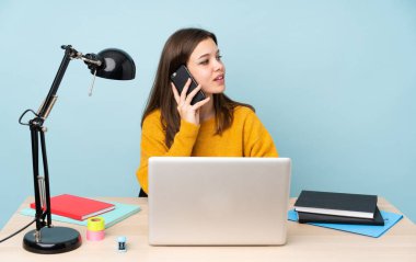 Öğrenci kız, mavi arka planda izole bir şekilde evinde çalışıyor cep telefonuyla sohbet ediyor.