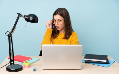 Evinde okuyan öğrenci kız mavi arka planda gözlüklü ve şaşırmış.