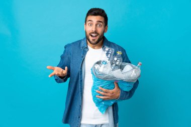 Kafkasyalı genç bir adam elinde plastik şişelerle dolu bir çanta tutuyor. Mavi arka planda izole edilmiş ve şok olmuş bir yüz ifadesi var.