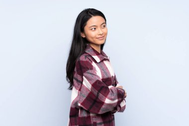 Genç Çinli bir kadın mavi arka planda izole edilmiş, yan tarafa bakıyor ve gülümsüyor.