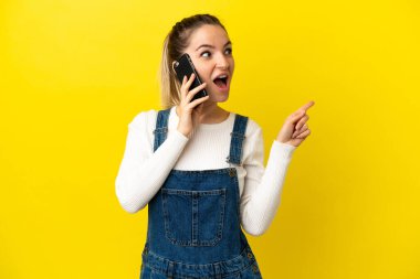 İzole edilmiş sarı arka plan üzerinde cep telefonu kullanan genç bir kadın parmak kaldırırken çözümü bulmayı umuyor.