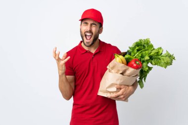 Sakallı genç adam elinde bir çanta dolusu sebzeyle beyaz arka planda izole edilmiş mutsuz ve hayal kırıklığına uğramış bir şekilde duruyor.