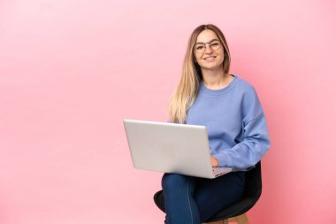 Dizüstü bilgisayarı olan genç bir kadın pembe arka plan üzerinde oturuyor ve kollarını ön tarafta çapraz pozisyonda tutuyor.