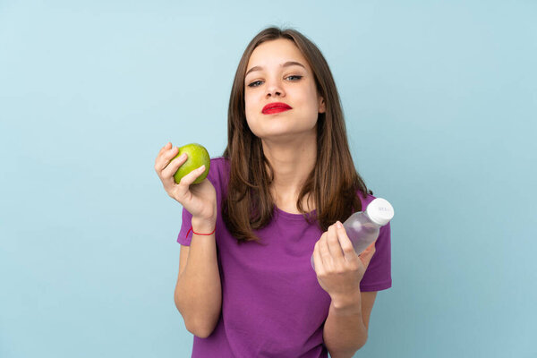 Teenager girl isolated on blue background with an apple and with a bottle of water