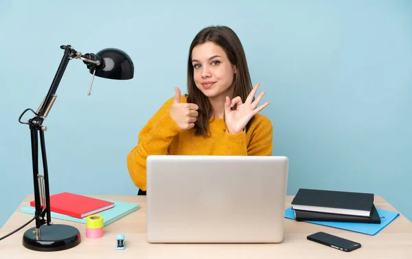 Student girl studying in her house isolated on blue background showing ok sign and thumb up gesture