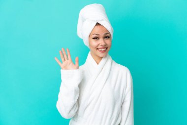 İzole edilmiş mavi arka planda bornozlu genç sarışın kız mutlu bir ifadeyle selamlıyor.