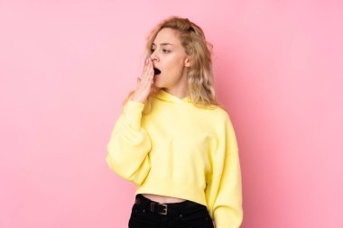 Pembe arka planda izole edilmiş bir sweatshirt giyen genç sarışın kadın esniyor ve ağzını eliyle kocaman açık tutuyor.