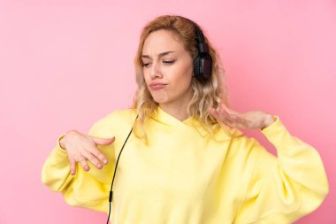 Pembe arka planda izole edilmiş sweatshirt giyen genç sarışın kadın müzik dinliyor ve dans ediyor.
