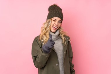 Kış şapkalı genç kız pembe arka planda izole edilmiş sana parmak atıyor.