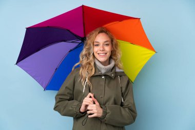 Mavi arka planda şemsiyesi olan genç sarışın kadın çok gülümsüyor.