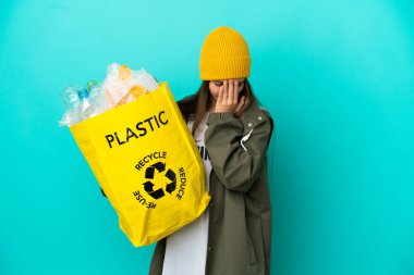 Elinde plastik şişelerle dolu bir çanta tutan küçük kız izole edilmiş mavi arka planda yorgun ve hasta bir ifadeyle geri dönüşüm yapıyor.