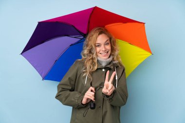 Mavi arka planda şemsiye tutan genç sarışın kadın gülümsüyor ve zafer işareti gösteriyor.