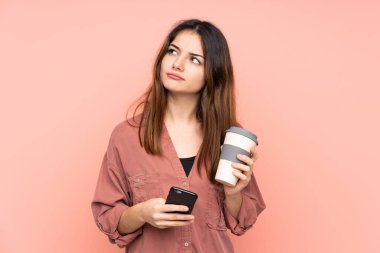 Kafkasyalı genç bir kadın pembe arka planda izole edilmiş. Elinde kahve ve bir şeyler düşünürken cep telefonu var.