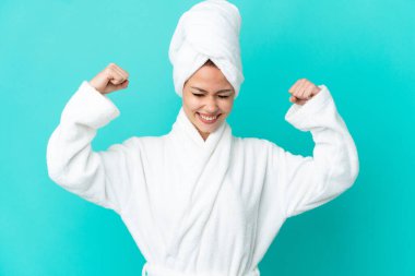 İzole edilmiş mavi arka planda bornozlu genç sarışın kız güçlü bir jest yapıyor.