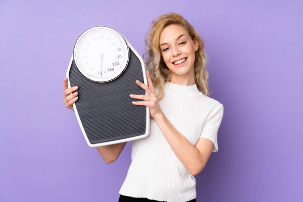 Young blonde woman isolated on purple background with weighing machine
