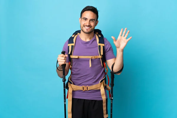 Jovem Caucasiano Com Mochila Postes Trekking Isolado Fundo Azul Saudando — Fotografia de Stock