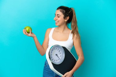 Mavi arka planda izole edilmiş beyaz bir kadın. Elma ararken elinde bir tartı makinesi tutuyor.