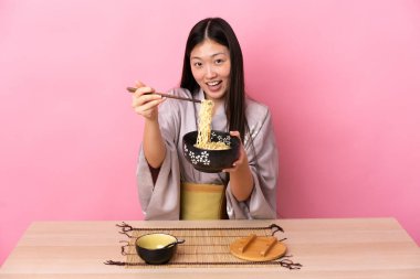 Genç Çinli kız kimono giyiyor ve pembe arka planda bir masada erişte yiyor.