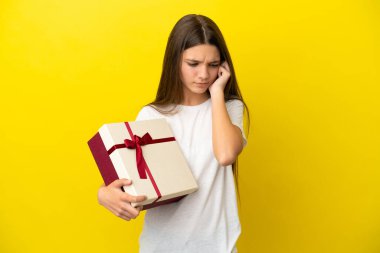 İzole edilmiş sarı arka planda hediye taşıyan küçük kız hayal kırıklığına uğramış ve kulaklarını kapatmış.