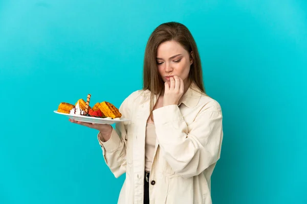 Adolescente Menina Segurando Waffles Sobre Isolado Fundo Azul Com Dúvidas — Fotografia de Stock