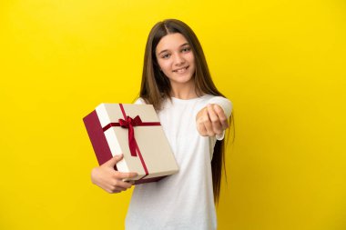 Küçük kız, izole edilmiş sarı arka planda bir hediye tutuyor. Para hareketi yapıyor.