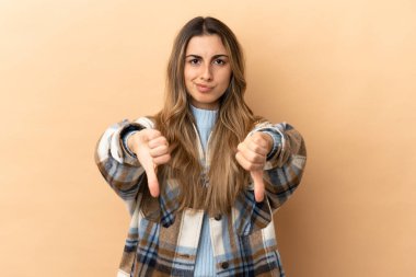 Beyazlı genç bir kadın bej arka planda izole edilmiş, baş parmağını iki eliyle aşağı gösteriyor.
