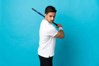 Genç yakışıklı adam mavi arka planda izole edilmiş tenis oynuyor.