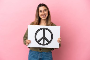 Pembe arka planda izole edilmiş beyaz bir kadın elinde mutlu bir ifadeyle barış sembolü taşıyan bir levha tutuyor.