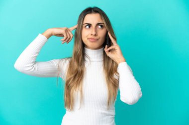 Mavi arka planda izole edilmiş cep telefonu kullanan beyaz bir kadın. Şüpheleri ve düşünceleri var.