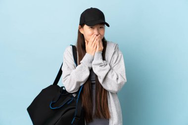 Spor çantalı genç Çinli kadın mavi arka planda mutlu ve gülümseyen elleri ve elleri ile ağzını kapatıyor.