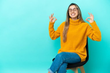 Sandalyede oturan beyaz bir kadın. Zen pozuyla mavi arka planda izole edilmiş.