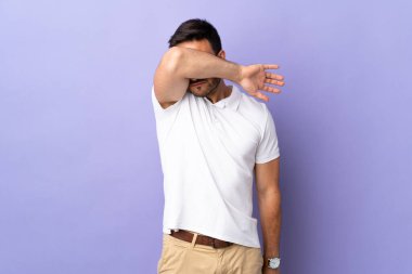 Mor arka planda izole edilmiş genç yakışıklı adam gözlerini elleriyle kapatıyor.